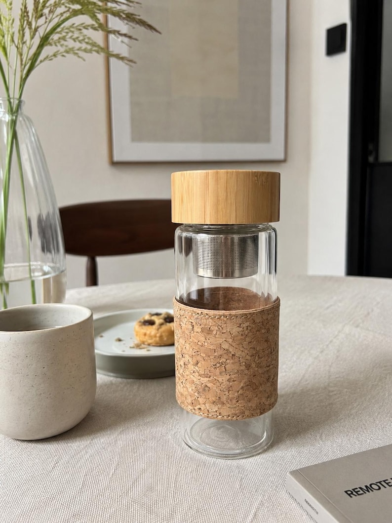 May include: A clear glass tea infuser bottle with a bamboo lid and a cork sleeve. The bottle has a stainless steel infuser basket inside. The bottle is sitting on a white tablecloth.