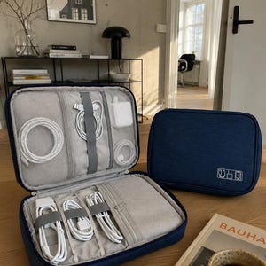 May include: Two navy blue rectangular electronic accessory organizers with zipper closures. One case is open, revealing interior mesh pockets, elastic straps, and a zippered compartment containing white charging cables and power adapters.