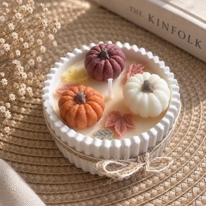 May include: A round white fluted candle container filled with wax, featuring three small pumpkin-shaped wax embeds in dark red, orange, and white, accented with miniature wax leaves and a single central wick, tied with a rustic twine bow.
