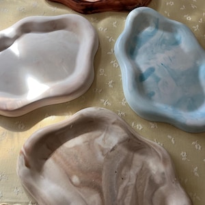 Op de afbeelding: Vier onregelmatige, golvende schalen met gladde oppervlakken. Eén schaal is donkerbruin met wervelende patronen, één is gebroken wit, één heeft een lichtblauw en wit marmer-effect en de vierde heeft een beige en wit gemarmerd ontwerp.