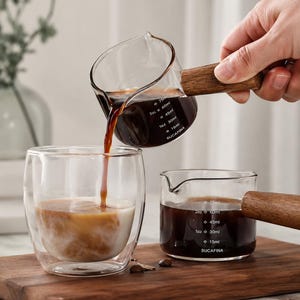 Könnte beinhalten: Ein Messbecher aus klarem Glas mit Holzgriff gießt dunkle Flüssigkeit in ein doppelwandiges Glas. Ein weiterer Messbecher mit Holzgriff steht auf einem Holzbrett. Der Text "SUCAFINA" ist sichtbar.