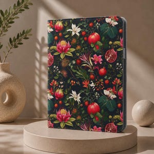 Könnte beinhalten: Eine rechteckige Tablethülle mit dunklem Hintergrund und einem dichten botanischen Muster aus roten Granatäpfeln, rosa Protea-Blüten, weißen Lilien und grünem Blattwerk. Das Etui hat ein schmales Profil mit grauem Rand und glatter Oberfläche.