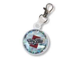 May include: A round keychain with a silver clasp and a white strap. The keychain has a holographic design with the words "Very Nice" and "YGO" in a stylized font. The text "FOR THE LOVE OF SHINY CARD BOARD" is printed around the edge.