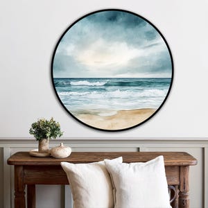 Könnte beinhalten: Ein rundes Wandkunstwerk mit einer Aquarellmalerei einer Meereslandschaft. Das Kunstwerk zeigt einen Strand mit schäumenden Wellen, einem sandigen Ufer und einem bewölkten Himmel. Das Gemälde ist mit einem schwarzen Rahmen versehen und schafft ein maritimes Thema.