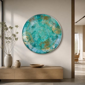 May include: A large circular wall art piece featuring a vibrant teal and turquoise watercolor-style background with metallic gold leaf accents and thin concentric circular line patterns, framed in a thin silver border mounted on a plain wall.