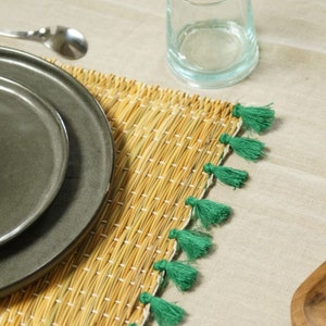 Op de afbeelding: Een geweven placemat in natuurlijke strokleur met witte stiksels. De placemat is afgewerkt met groene kwastjes. Een grijze plaat en een helder glas staan op de placemat, met een lepel op de achtergrond. De scène suggereert een eetsituatie.
