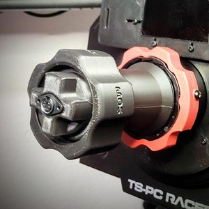 以下が含まれることがあります： 黒と赤の3Dプリントされたステアリングホイールアダプターで、側面に「TS-PC RACER」の文字が印刷されています。アダプターには、小さな銀色のボタンが付いた黒いノブがあります。
