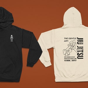 May include: Two hooded sweatshirts, one black and one cream. The black sweatshirt has a small skeleton graphic on the front. The cream sweatshirt has a graphic of two people in a jiu-jitsu hold with the text "The Gentle Art: Jiu Jitsu" and "Until someone taps out, passes out or snaps a limb. Ya know... gently."