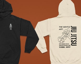 May include: Two hooded sweatshirts, one black and one cream. The black sweatshirt has a small skeleton graphic on the front. The cream sweatshirt has a graphic of two people in a jiu-jitsu hold with the text "The Gentle Art: Jiu Jitsu" and "Until someone taps out, passes out or snaps a limb. Ya know... gently."