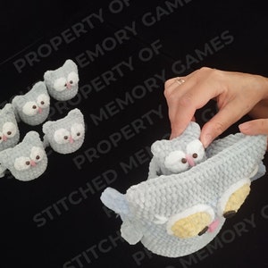 May include: A set of gray crocheted owl toys. The owls have black eyes, white pupils, and pink beaks. One owl is larger than the others and has yellow accents around the eyes. The owls are arranged on a black surface.