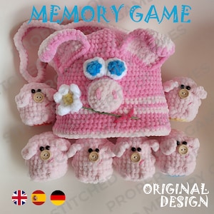 May include: A pink crocheted pig memory game with a flower and a green stem. The pig has blue eyes and a pink snout. There are six smaller pink crocheted pigs with brown buttons for eyes. The text "MEMORY GAME" is written in blue letters at the top of the image. The text "ORIGINAL DESIGN" is written in white letters on a blue background at the bottom of the image.