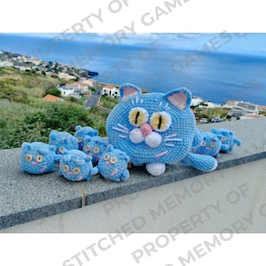 May include: A blue crocheted cat with yellow eyes and a pink nose, surrounded by seven smaller blue crocheted cats with yellow eyes. The cats are sitting on a gray surface.
