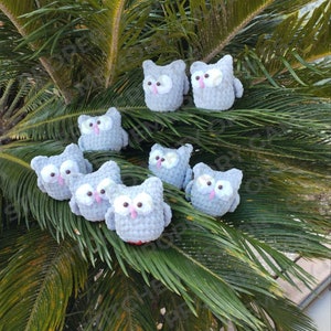 May include: A collection of gray crocheted owl toys with pink eyes and beaks. The owls are arranged on a green palm frond.