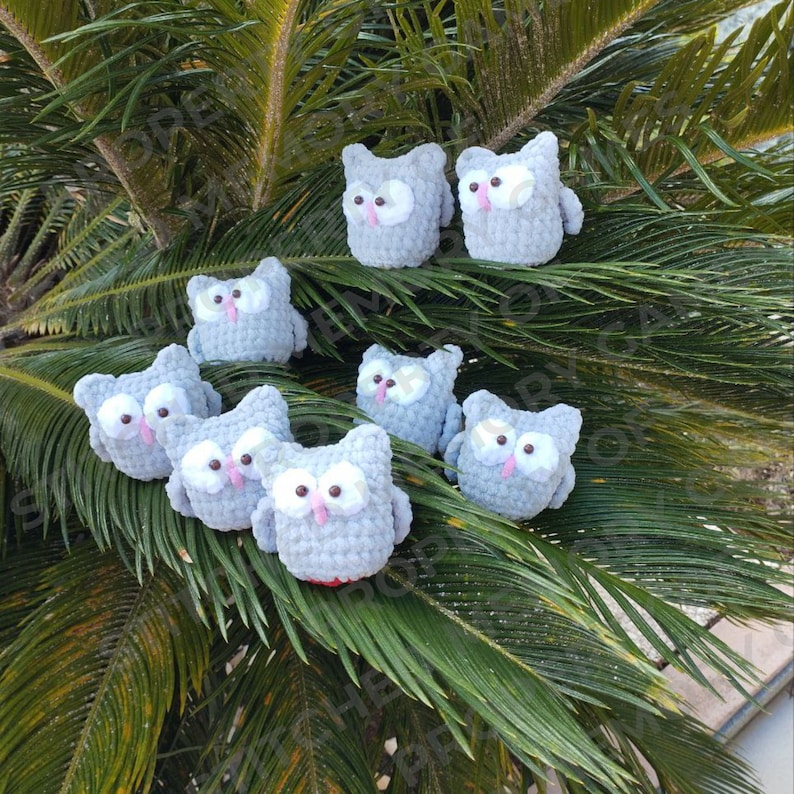May include: A collection of gray crocheted owl toys with pink eyes and beaks. The owls are arranged on a green palm frond.