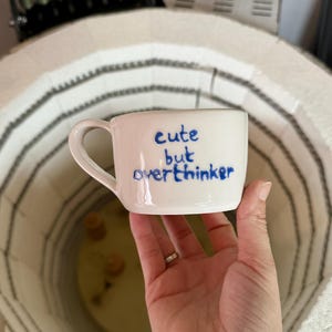 Puede incluir: Una taza de cerámica blanca con un asa curva, con el texto azul "cute but overthinker". La taza se sostiene en una mano, con un horno visible en el fondo. La taza es de tamaño estándar para café o té.