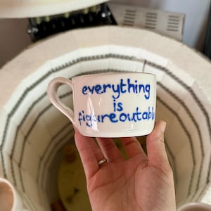 May include: A white ceramic mug with a black handle and the text "But First, Coffee" in a bold, black font. The mug is set against a blurred background, suggesting a cozy setting.