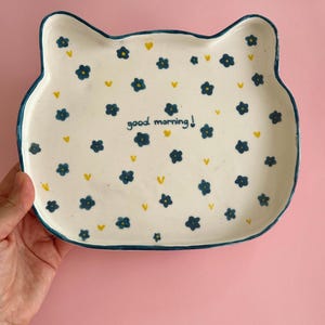 Può includere: Un piatto in ceramica a forma di gatto con base color crema, caratterizzato da un motivo di piccoli fiori blu scuro e minuscoli cuori gialli. Il testo "good morning!" è stampato in blu scuro al centro e il bordo è delineato in blu scuro.