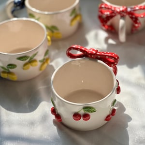 May include: White ceramic mugs with decorative fruit and bow accents. One mug features red cherries, another has yellow lemons, and a third has a red polka dot bow. The mugs have a cream-colored interior and are set on a white surface.
