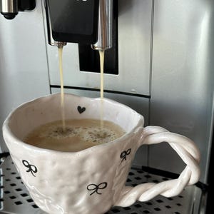 Może przedstawiać: Biały ceramiczny kubek ze skręconym uchwytem jest napełniany kawą z ekspresu do kawy Philips. Kubek jest ozdobiony czarnymi kokardkami i sercami. Ekspres jest srebrny i czarny.