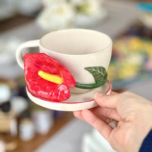 May include: Cream-colored ceramic teacup and saucer set with a red anthurium flower design. The flower has a yellow spadix. The saucer and cup have a red trim. A green leaf is painted on the cup. The cup is held by a person.