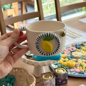 May include: A white ceramic mug with a lemon design and a small bee. The mug has a blue and white striped handle and a lemon-shaped design on the front. The mug is held in a hand.