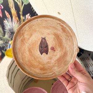 May include: A handmade ceramic plate featuring a brown bear illustration. The plate has a speckled, earthy tone with a brown bear and pink floral accents. The plate is held in a person's hand, with two other plates visible below.
