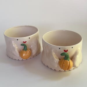Puede incluir: Dos cuencos de cerámica blanquecina con forma de fantasma y detalles de calabaza naranja. Cada cuenco tiene un ojo de punto negro y una boca curva roja. Las calabazas tienen tallos verdes. Los cuencos son para decoración de Halloween o otoño.