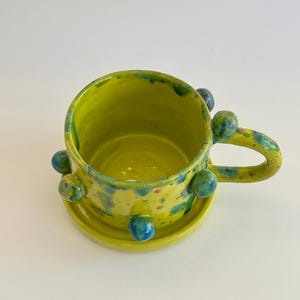 Puede incluir: Una taza de cerámica de color verde lima brillante con su plato a juego, con un acabado brillante y motas azules y amarillas. La taza está decorada con varios bultos redondeados en relieve en el exterior e incluye un asa curva.