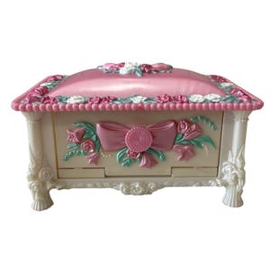 May include: A decorative jewelry box in a pastel color palette. The box features a pink, cushioned top with floral accents and a bow design on the front. The box is supported by ornate legs.