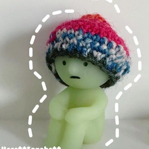 Puede incluir: Una pequeña figurita verde con una expresión triste que lleva un gorro de punto multicolor. La figurita está sentada con las piernas cruzadas.