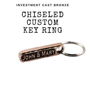 May include: A bronze key ring with the text "JOHN & MARY" engraved on a rectangular tag. The tag is attached to a silver-colored ring. The text "INVESTMENT CAST BRONZE" and "CHISELED CUSTOM KEY RING" are printed above the key ring.
