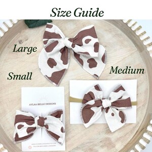 May include: A size guide displays three fabric hair bows in brown and white cow print. The bows are labeled as Large, Medium, and Small. The Small bow is attached to a card with the text "AYLAA BELLE DESIGNS". The bows are on a wooden tray.