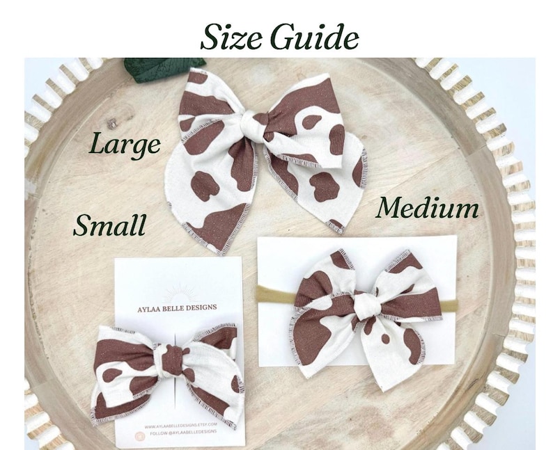 May include: A size guide displays three fabric hair bows in brown and white cow print. The bows are labeled as Large, Medium, and Small. The Small bow is attached to a card with the text "AYLAA BELLE DESIGNS". The bows are on a wooden tray.