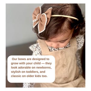 May include: A brown bow headband with a white trim is worn by a child. The child is wearing a tan overall dress and a floral print shirt. The text in the image says the bows are designed to grow with your child.