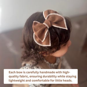 May include: A brown fabric bow with white stitching detail is worn in the hair. The bow is a large, decorative accessory. The text in the image says: Each bow is carefully handmade with high-quality fabric, ensuring durability while staying lightweight and comfortable for little heads.