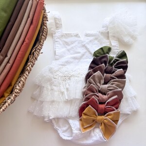 May include: A white baby romper with a layered tulle skirt and a collection of colorful fabric bows. The bows are in shades of green, brown, tan, and mustard yellow. A basket of folded fabric in various colors is in the background.