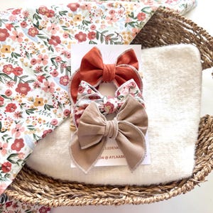May include: A woven basket holds a white plush blanket, a floral patterned blanket, and three fabric hair bows. The bows are in shades of rust, floral print, and tan. The floral blanket has a white background with pink and red flowers.