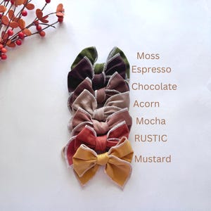 May include: A stack of seven velvet hair bows in various colors, including moss, espresso, chocolate, acorn, mocha, rustic, and mustard. Each bow has a contrasting trim and is displayed on a white background. The names of the colors are listed vertically.