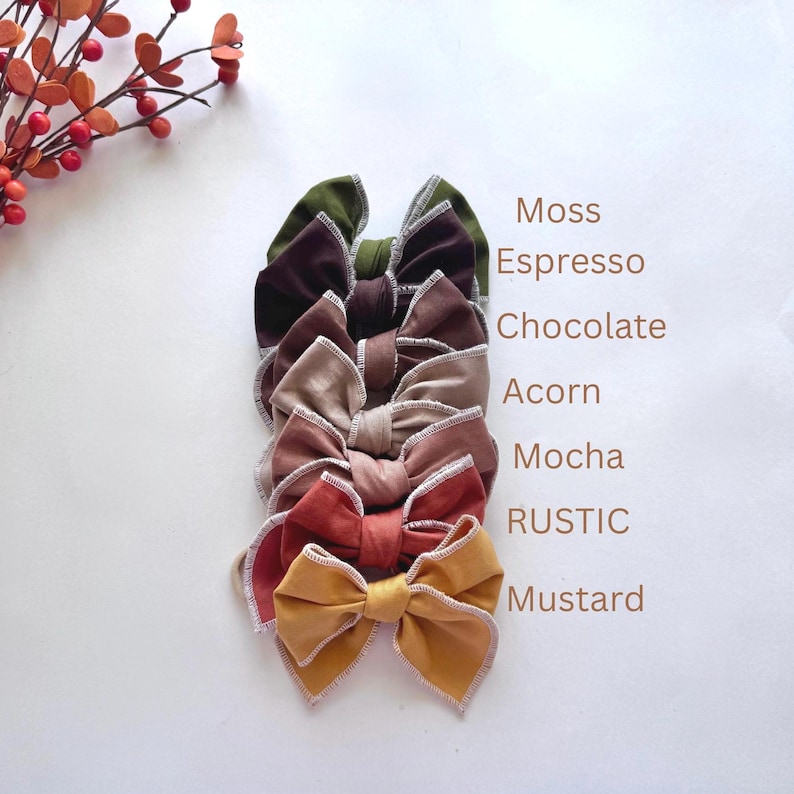 May include: A stack of seven velvet hair bows in various colors, including moss, espresso, chocolate, acorn, mocha, rustic, and mustard. Each bow has a contrasting trim and is displayed on a white background. The names of the colors are listed vertically.