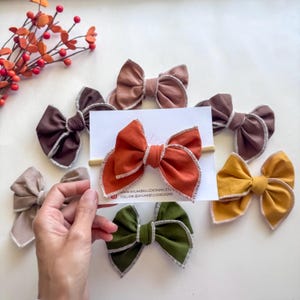 May include: A collection of fabric hair bows in various fall colors, including rust orange, olive green, and mustard yellow. Each bow features a contrasting white stitched edge. A hand holds a bow on a headband. The bows are arranged on a white surface.