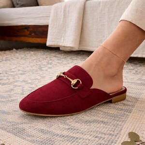 Könnte beinhalten: Burgunderfarbener Wildleder-Slipper mit goldfarbenem Metall-Trensen-Detail. Der Schuh hat eine runde Spitze und einen niedrigen Absatz. Der Schuh steht auf einem gemusterten Teppich.