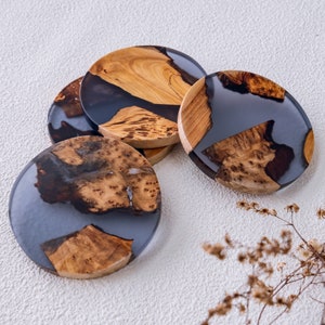 Könnte beinhalten: Eine Sammlung handgefertigter runder Untersetzer. Jeder Untersetzer hat ein einzigartiges Design mit natürlichen Holzmaserungsmustern, die in einem durchscheinenden, dunkelgrauen Harz eingeschlossen sind. Die Untersetzer bestehen aus Holz und Harz.