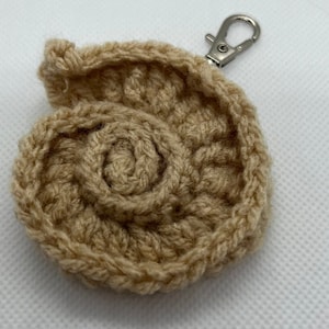 May include: A beige crocheted keychain shaped like a spiral shell with a silver metal key ring.