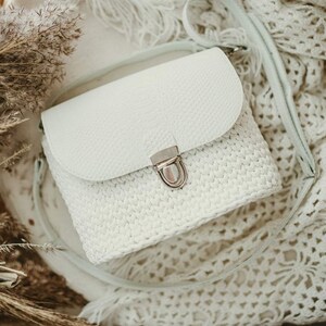 Puede incluir: Un bolso bandolera blanco con una solapa texturizada de color sólido y un cuerpo de ganchillo. El bolso incluye un cierre de presión de metal plateado y una correa de hombro blanca a juego, ofreciendo un aspecto estructurado y suave.