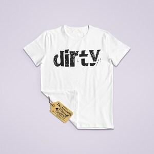 May include: White t-shirt with the word "dirty" printed in black, distressed font across the chest. A small tag with text is attached to the shirt. The shirt is displayed on a light purple background.