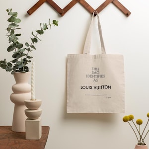 May include: A beige tote bag with the text "THIS BAG IDENTIFIES AS LOUIS VUITTON" hangs from a wooden wall rack. A pink vase with eucalyptus and a candle sits on a wooden table. Another pink vase with yellow flowers is also visible.