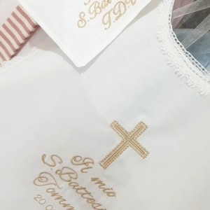 Può includere: Fazzoletto da battesimo bianco con croce ricamata in oro e testo. Il testo recita "Il mio S. Battesimo Tommaso 20.04.2025" e "Il mio S. Battesimo T.D.V.". Il fazzoletto ha un bordo in pizzo e un dettaglio in tessuto a righe.
