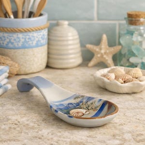 May include: A ceramic spoon rest featuring a hand-painted coastal scene with blue brushstrokes, green seagrass, and a detailed seashell motif. The spoon rest has a curved handle and a smooth, glossy finish, designed to hold kitchen utensils.