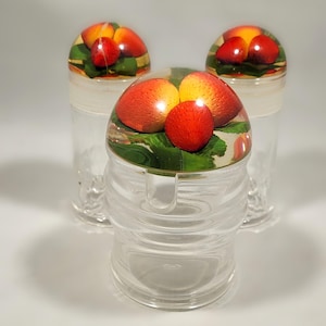 May include: Three clear glass jars with lids featuring a colorful fruit design. The lids are round and have a raised design of strawberries on a green leaf background.