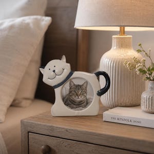 May include: A cream-colored ceramic cat-shaped picture frame with a dark blue collar and tail. The frame features a circular opening displaying a photograph of a tabby cat. The cat figure has a smiling face with black eyes and a nose.
